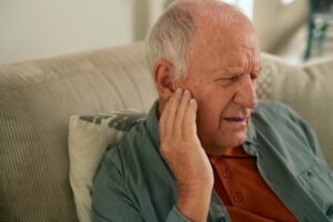 Senior man holding ear in pain while sitting on couch, showing tinnitus, hearing loss, and ear discomfort.
