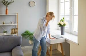 Weary woman standing by window with hand on forehead, feeling dizzy and exhausted, concept of vertigo, fatigue, or heat discomfort.