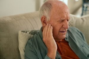 Senior man holding ear in pain while sitting on couch, showing tinnitus, hearing loss, and ear discomfort.